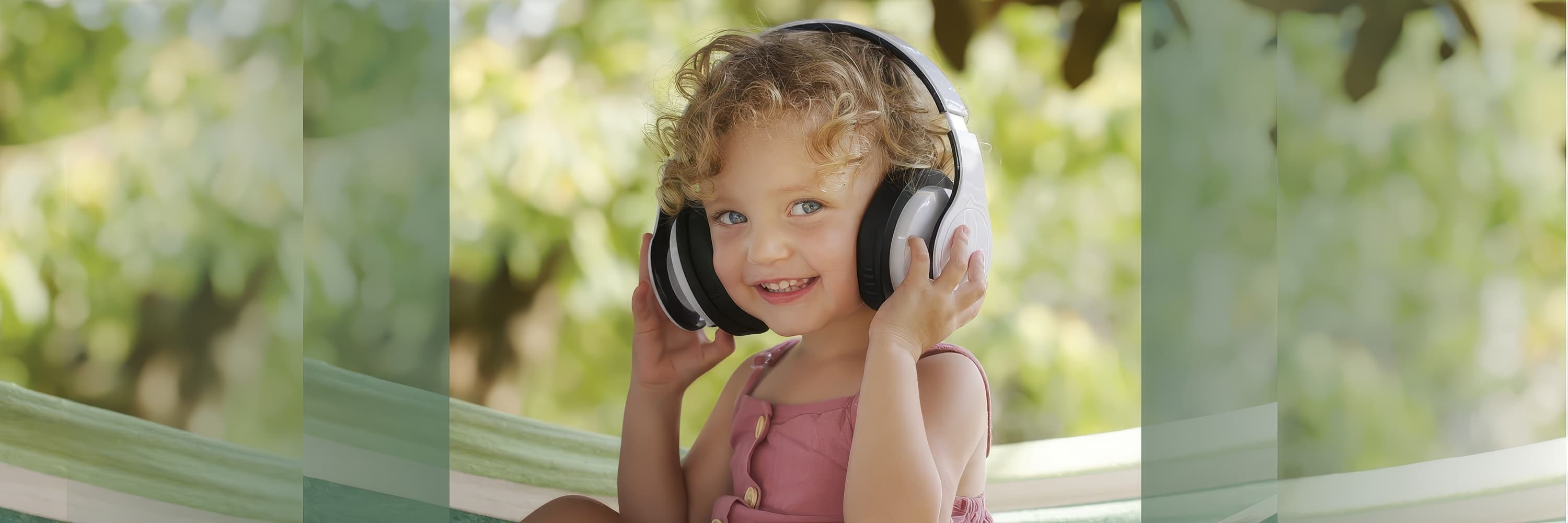 Smiling child in a pink dress wearing large headphones while sitting outdoors