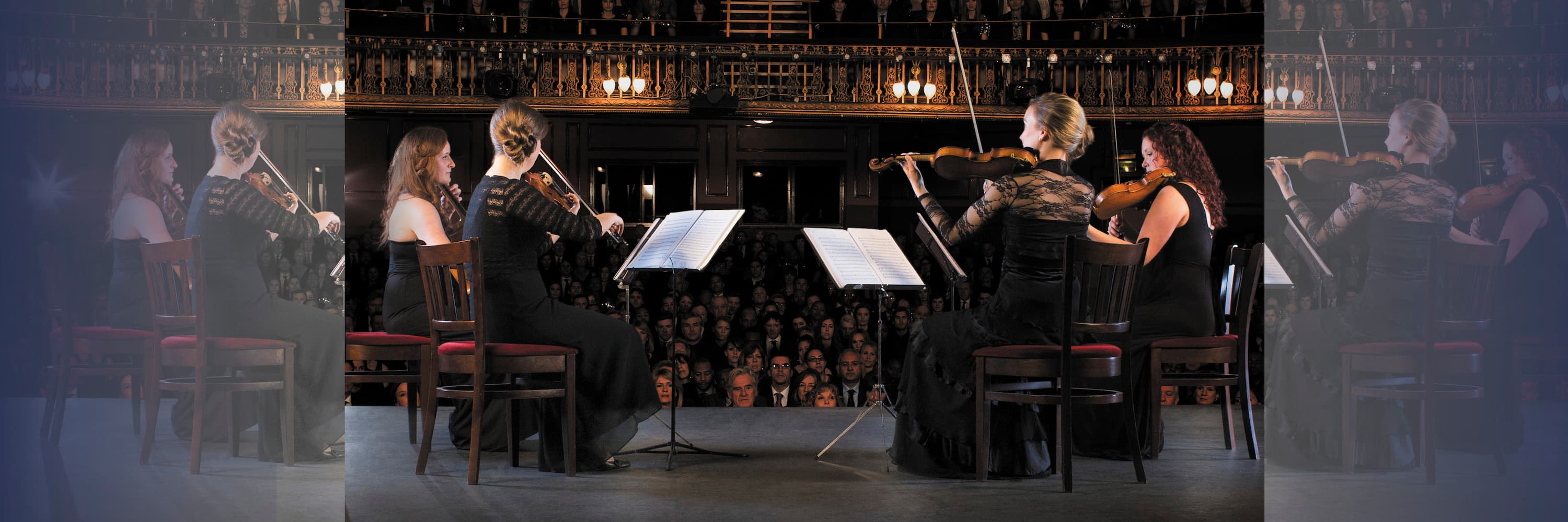 Quartet of female violinists performing on stage in front of a large seated audience.