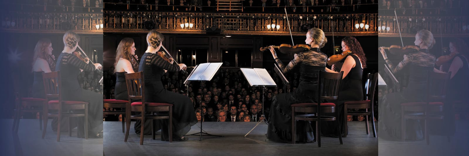 Quartet of female violinists performing on stage in front of a large seated audience.