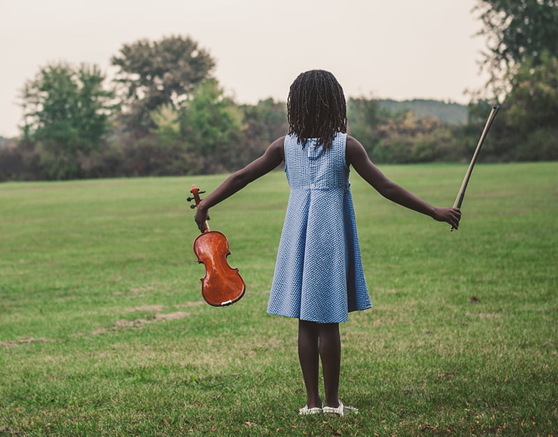 Young girl in a blue dress holding a violin and bow in an open field.