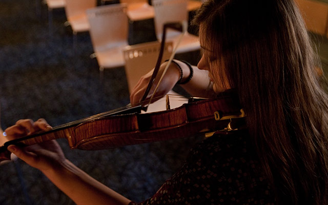 Woman playing violin on stage with empty chairs in the background.