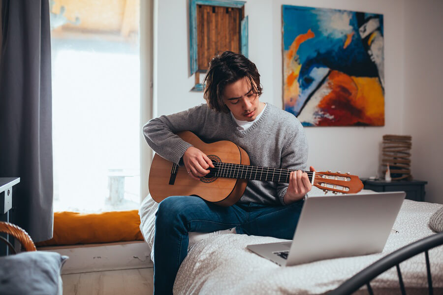 Young man playing guitar on a bed with a laptop in front of him