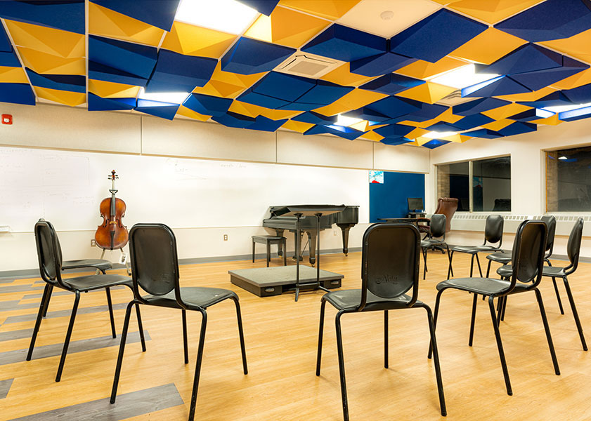 Music classroom with chairs arranged in a circle, a piano, a cello on a stand, and a colorful blue and yellow ceiling design.