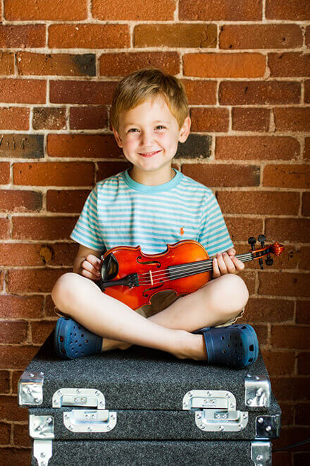 A youg boy sitting against a brick wall with a violin on his lap