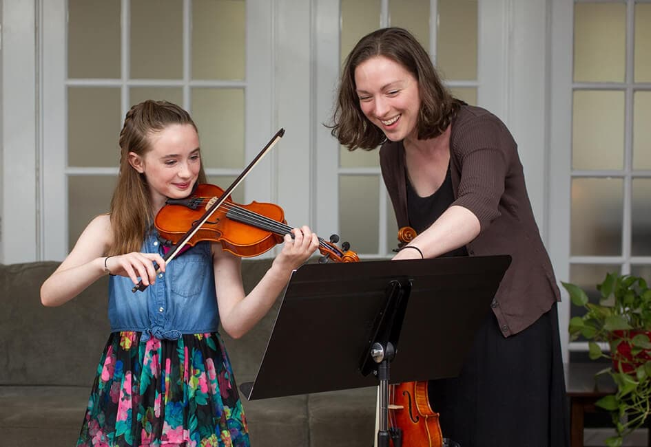 Woman points at sheet music on a stand while girl plays the violin