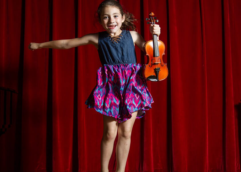 Girl mid-jump while holding a violin in front of a maroon theatre curtain