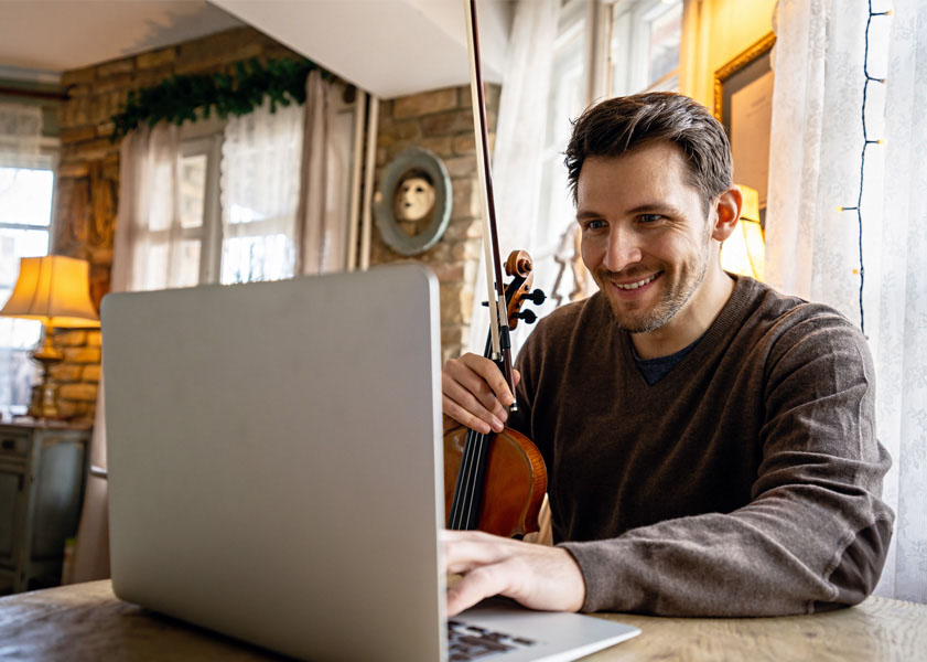 Man smiling while holding a violin and using a laptop in a cozy, well-lit room with rustic decor.
